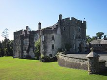 Grey walled castle of three and four storeys partly in its own shadow on a sunny day with bright blue sky and green lawns to foreground