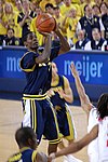 Pemain basket biru gelap seragam menembak jump shot di atas outstreched lengan bek putih.