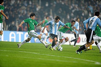 Fotografie fotbalového zápasu, jeden hráč v modré a bílé běží s míčem sledovaným dvěma hráči v zelené barvě