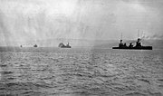 A line of four ships underway: the rightmost ship is the closest to the camera, while the leftmost ship is very distant. A landmass stretches along the horizon, behind the ships.