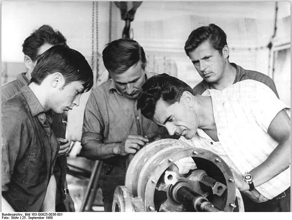 A master discusses a vacuum compressor with his apprentice boy and several other craftsmen.