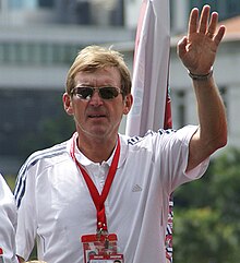 Colour photograph of Dalglish in Singapore, 2009. He is dressed casually, wearing sunglasses and has his hand raised
