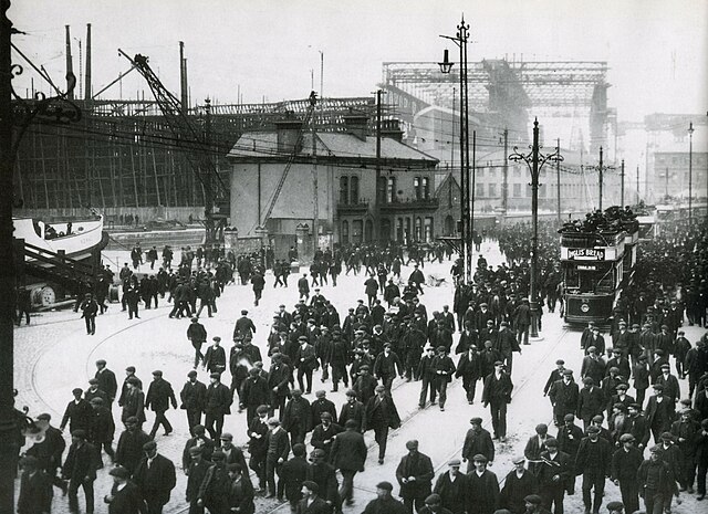 Workers leaving the shipyard at Queens Road in early 1911. RMS Titanic is in the background, beneath the Arrol Gantry. The bow of SS Nomadic is at the