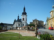 Fotografie náměstí. V popředí je postavena zídka s barokními sochami. V pozadí je pak bílý kostel.