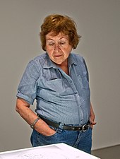 Teresa Burga, de 77 anos, está atrás de uma mesa e, com as mãos nos bolsos da calça, olha um desenho.