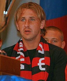 A white blond male seating on a stand with a black and red FC Khimki scarf around his neck.