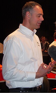Photo de profil droit Nick Wirth en 2011, vêtu d'une chemise blanche , devant une voiture rouge