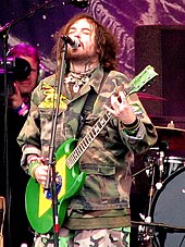 A man in his thirties, singing into a microphone while playing guitar.