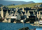 Stone houses crowd around a shore, the gable ends facing the water, with green hills beyond.