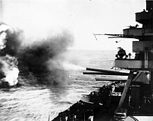 Seen from the side of a ship as large guns fire toward targets out of sight