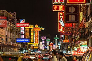 A street during sunset lined with many stalls and shops with a lot of signs bearing Thai and Chinese names