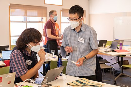 Wearing face masks two people are smiling and looking at bits of paper. One is sitting at a table and one is standing. Other people are in the background out of focus.