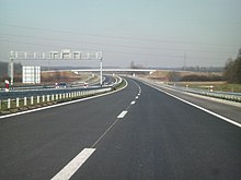 Une vue de l'autoroute à quatre voies avec un terre-plein central, prise depuis un niveau de chaussée.  L'arrière d'un portique portant des panneaux de signalisation variables est visible sur la chaussée de gauche.