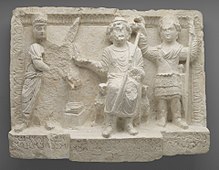 a relief depicting three figures. In the middle a deity sitting on a throne, to the right, a man (king Seleucus I) standing, and to the left a Palmyrene merchant is standing