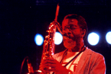 un saxophoniste avec barbe et lunettes sous l'éclairage rouge de la scène