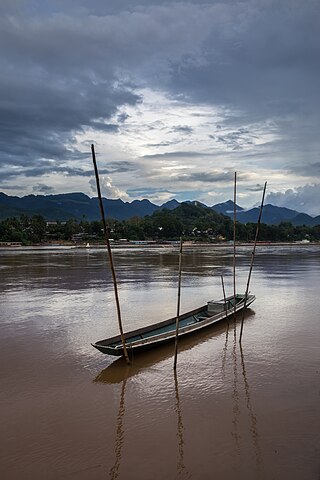 Pirogue moored to the Mekong bank with bamboo stakes under clearing of a sky reflected in muddy water