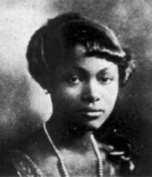 A young Black woman with hair cut in a short fringe, wearing a strand of pearls
