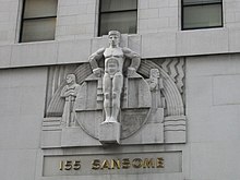 A color photograph of a nude male figure carved in stone over the entrance of a commercial building. The figure stands straight with arms akimbo and hands atop a stone disc behind the figure, flanked by geometric designs and two smaller figures of workmen looking off to each side. Below the sculpture grouping, raised gold letters read "155 Sansome".