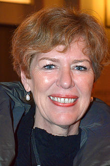 A close-up photo of a white woman with blue eyes and short red hair. She is dressed in black and has earrings on.