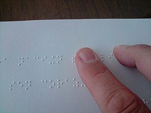 Zeige- und Mittelfinger eines Lesenden gleiten über die obere von zwei Zeilen Braille-Text. Deutlich zu erkennen sind die punktförmigen Erhöhungen in dem weißen Papier, die die Schriftzeichen bilden.