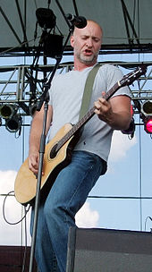 Full length portrait, wearing gray t-shirt and blue jeans, no hair, short gray beard, playing electric guitar.