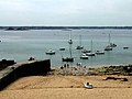 Douze bateaux sur l'eau calme ; à gauche, le débarcadère en béton. Au premier plan, la plage avec une petite embarcation sur le sable.