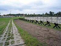 Strip of bare ploughed earth flanked by a concrete road on one side and a row of barricades and a fence on the other side, with buildings visible in the far background.