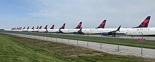 Muchos aviones en línea con el logo de Delta Air Lines en la cola, estacionados en el pavimento detrás de una valla.