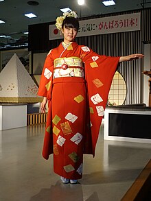 A young unmarried Japanese woman wearing a deep orange furisode with her arms outstretched.