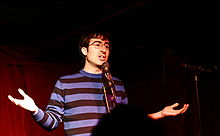Photo of Oliver wearing a black and blue striped shirt, performing standup in front of a red curtain