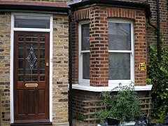 A brown building. Visible is a door at left, with a bowfront window at right; various tomato plants are also visible growing in front of the window.