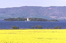 Поле желтых цветов на переднем плане и темно-синее озеро за ним. Лесистый остров в озере имеет белое двухэтажное здание в центре, а за ним - зеленые и коричневые холмы. Справа на дальнем склоне холма - небольшая группа домов.
