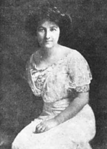 A white woman with dark hair in an updo, seated, wearing a light-colored blouse and skirt