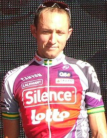 Seorang pria di usia dua puluhan yang mengenakan warna merah, ungu, dan putih bersepeda jersey, dengan hijau dan emas arm band.
