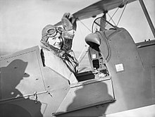 Une femme, en costume d’aviatrice adapté au froid, salue de la main dans un cockpit d’avion.