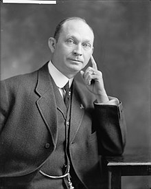 A balding man wearing a black jacket, vest, and tie, and white shirt, seated, leaning his left elbow on a table and his face against his extended left index finger