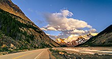 Sunwapta river in the foreground and Athabasca mountain the background. Asphalt road in the mountains, near the river with rocks and rocks, coniferous trees, snow-capped mountains in the background, lush clouds in the blue sky.