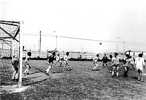 תמונה בשחור-לבן. משחק כדורגל. השער בשמאל התמונה, השוער עומד בשער מביט לימין התמונה, שם מספר חברי הקבוצות המשחקות, כולם מביטים לעבר כדור המשחק העף באוויר.