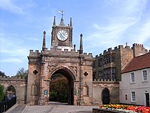 A crenellated stone arched gateway. In the middle is a small tower with a blue clock