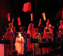 Image d'une chanteuse brune habillée en rouge, entourée par des cuivres et des bagnères