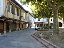 Fotografía en color de casas con entramado de madera con voladizo en la planta baja.  La plaza está sombreada por plátanos.