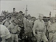 A group of men wearing military uniforms with a ship in the background