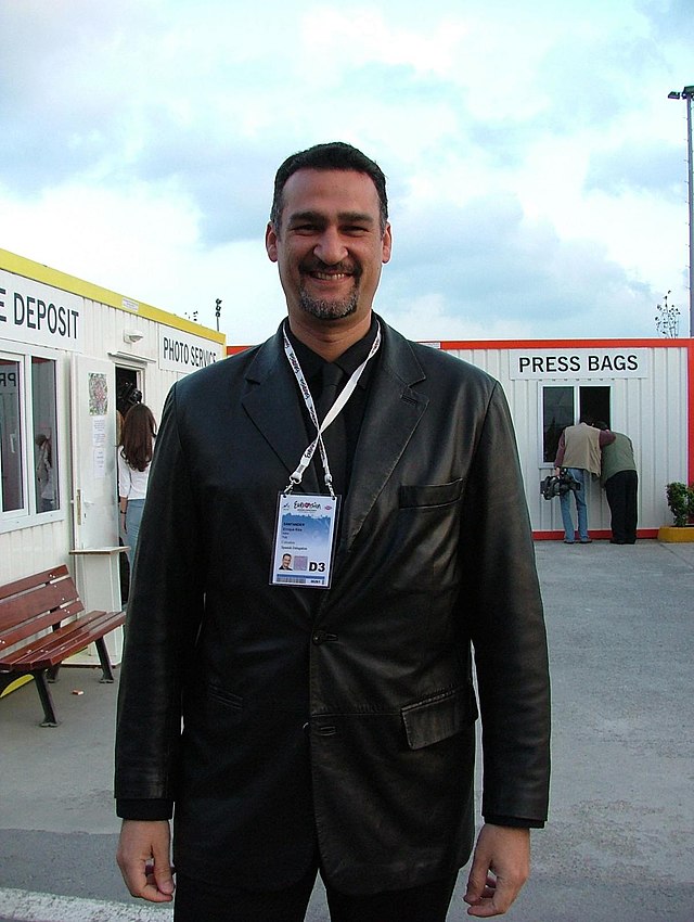 A man standing in a white shirt and his arms in his pockets leaning against a white pillar.