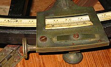 Details on an octant. This photo shows the graduated scale and the end of the index arm with the vernier. The thumbscrew used to lock the index arm position is seen below the index arm while the thumbscrew used for fine adjustment of the arm is on the left. To the right of the value 50 on the main scale, the SBR logo is engraved. The scale is directly graduated in degrees and thirds of a degree (20'). The vernier can divide the 20' intervals to the nearest minute of arc. Octant-scale-and-index.jpg