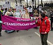 Olena Schewtschenko ist in einer roten Jacke und mit einem Megafon auf einer Demonstration zu sehen. Im Hintergrund sind viele andere Menschen, die ein pinkfarbenes Transparent halten.