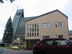 View from the west of Pielisensuu Church shows the steeply pyramidal church tower on the left, and roof line sloping inwards to the left over the parish hall and offices at the picture's right.