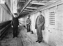 A prison corridor with a white man in uniform, a blurred white man in another uniform and a white man in a suit with his hands in his pockets.