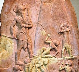 Zdjęcie fragmentu steli wyrzeźbionej w płaskorzeźbie.  Widzimy po lewej dużą stojącą postać w hełmie z dwoma rogami.  Idzie na prawo, gdzie znajdują się cztery małe upadłe lub błagalne postacie.  Powyżej wygrawerowany jest napis pismem klinowym.