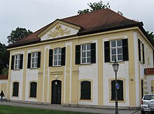 A square, two-storey building, painted in alternating yellow and white, with a decorative gable and a brick roof.