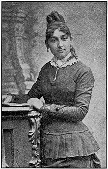 A young Black woman with hair in an elaborate updo, wearing a corseted dress or suit with a pleated peplum, standing in a portrait studio